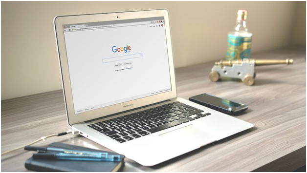 A laptop displaying the Google homepage sits on a desk next to a smartphone, a notebook with a pen, a bottle, and a small decorative cannon.